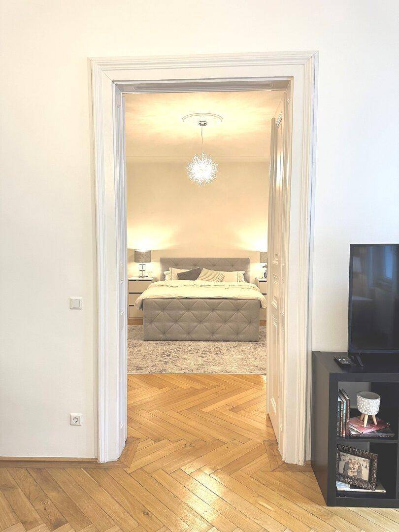 Blick durch eine offene Tür in ein helles Schlafzimmer mit großem Bett und Parkettboden.