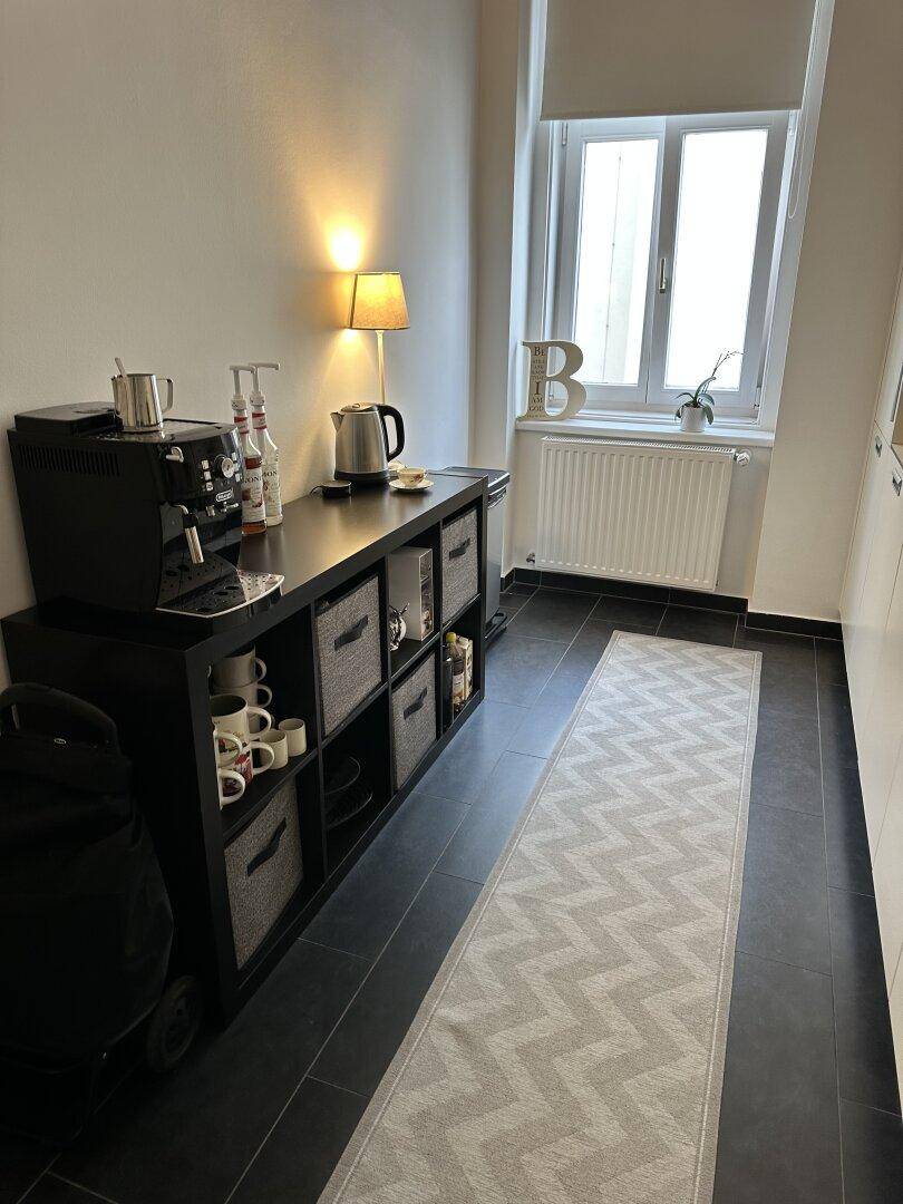 Küchenzeile mit Kaffeestation, Stauraum und Fenster, ergänzt durch dunkle Bodenfliesen.
