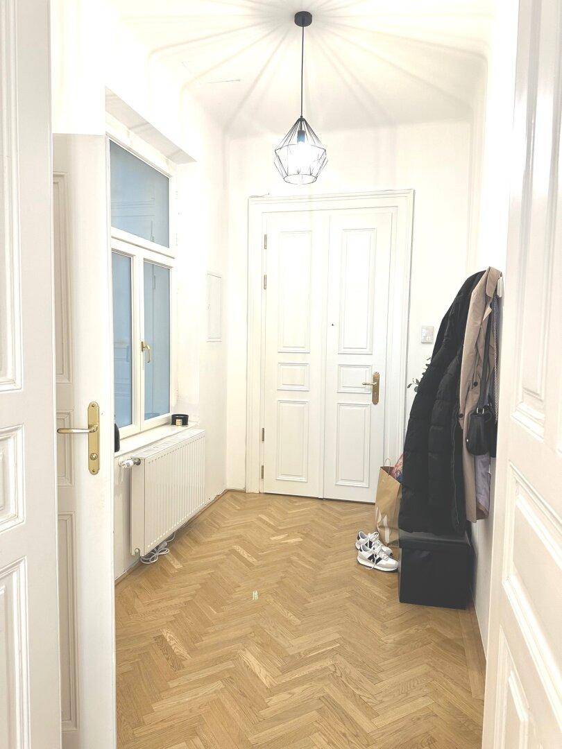 Heller Flur mit Fischgrätparkett, hohen Türen und einem Fenster für Tageslicht.