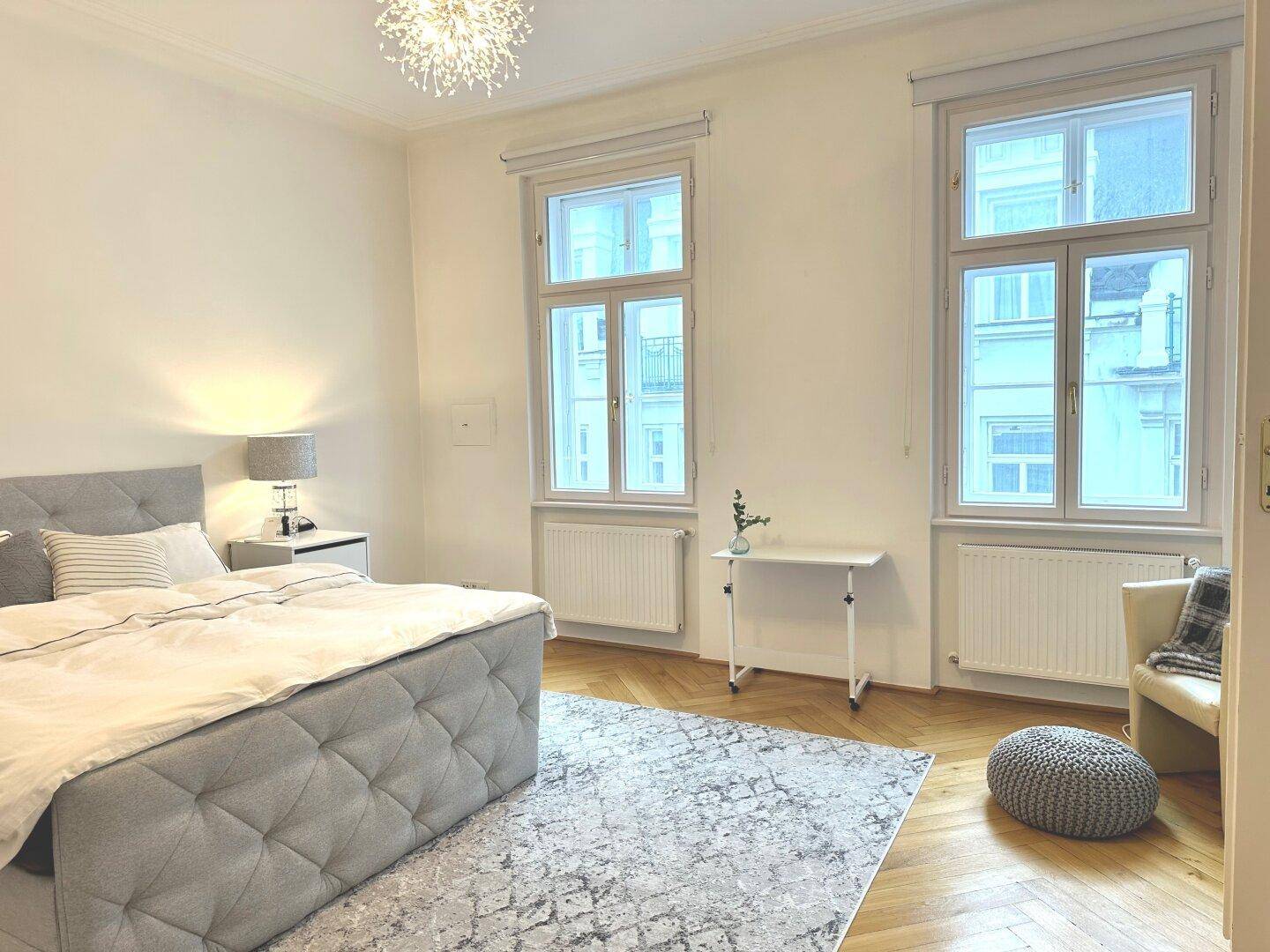 Geräumiges Schlafzimmer mit Fischgrätparkett, großem Bett und zwei hohen Fenstern.