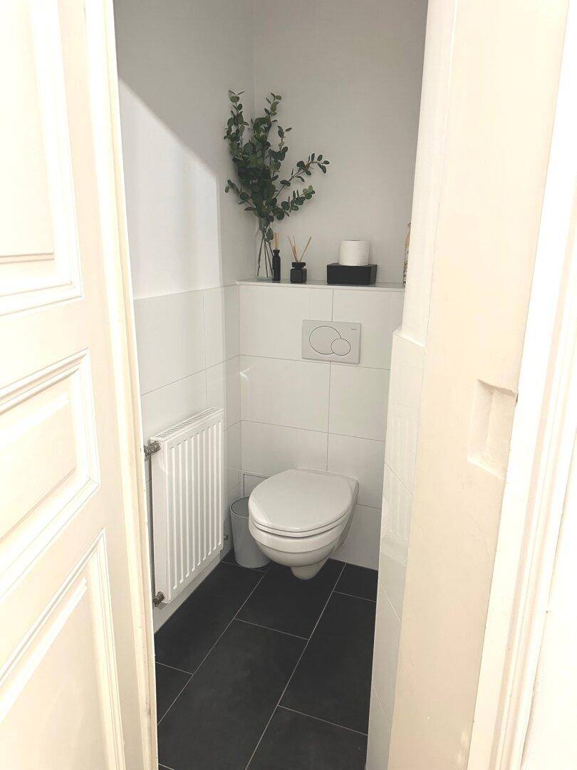 Separates WC mit Wand-WC, weißen Fliesen und Heizkörper.