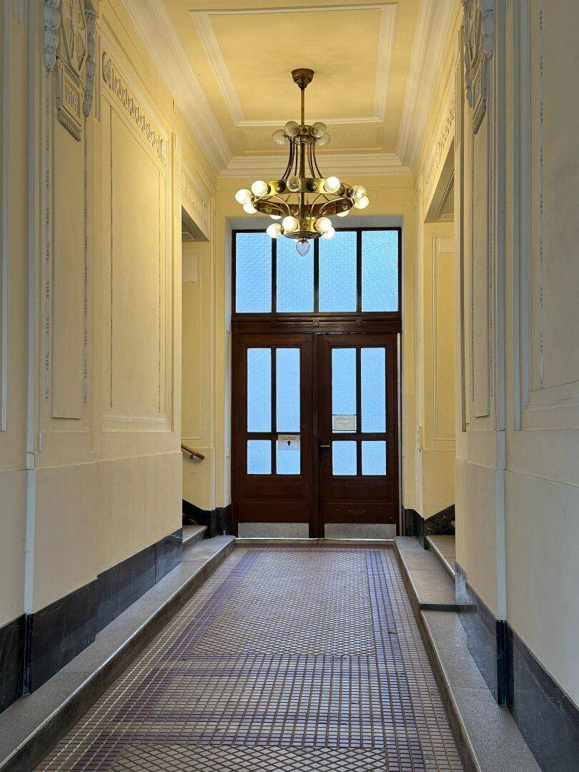 Langer, eleganter Flur mit Kronleuchter und hohen Doppeltüren, die Tageslicht hereinlassen.