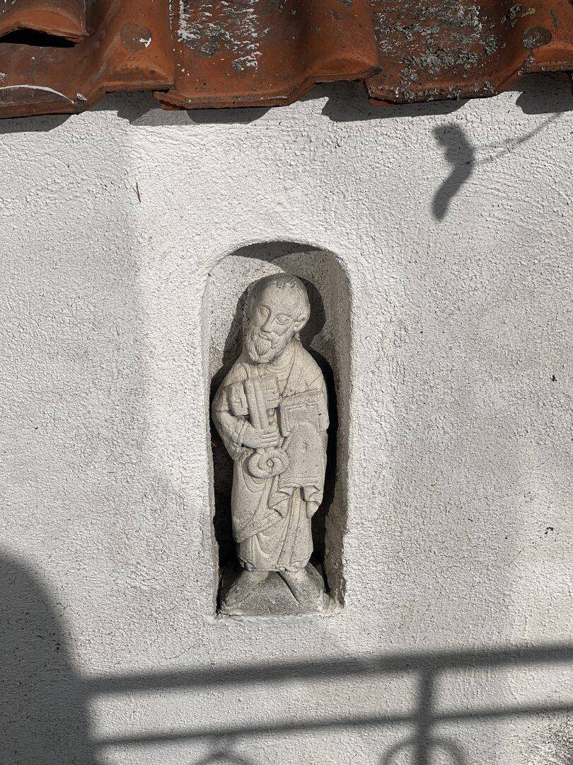 Eine steinerne Statue in einer Nische schmückt die weiße Außenwand unter einem Ziegeldach.
