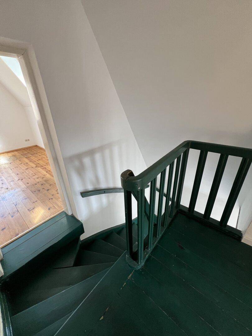 Grün gestrichene Holztreppe führt in einen hellen Raum mit Holzdielenboden und Dachschräge.