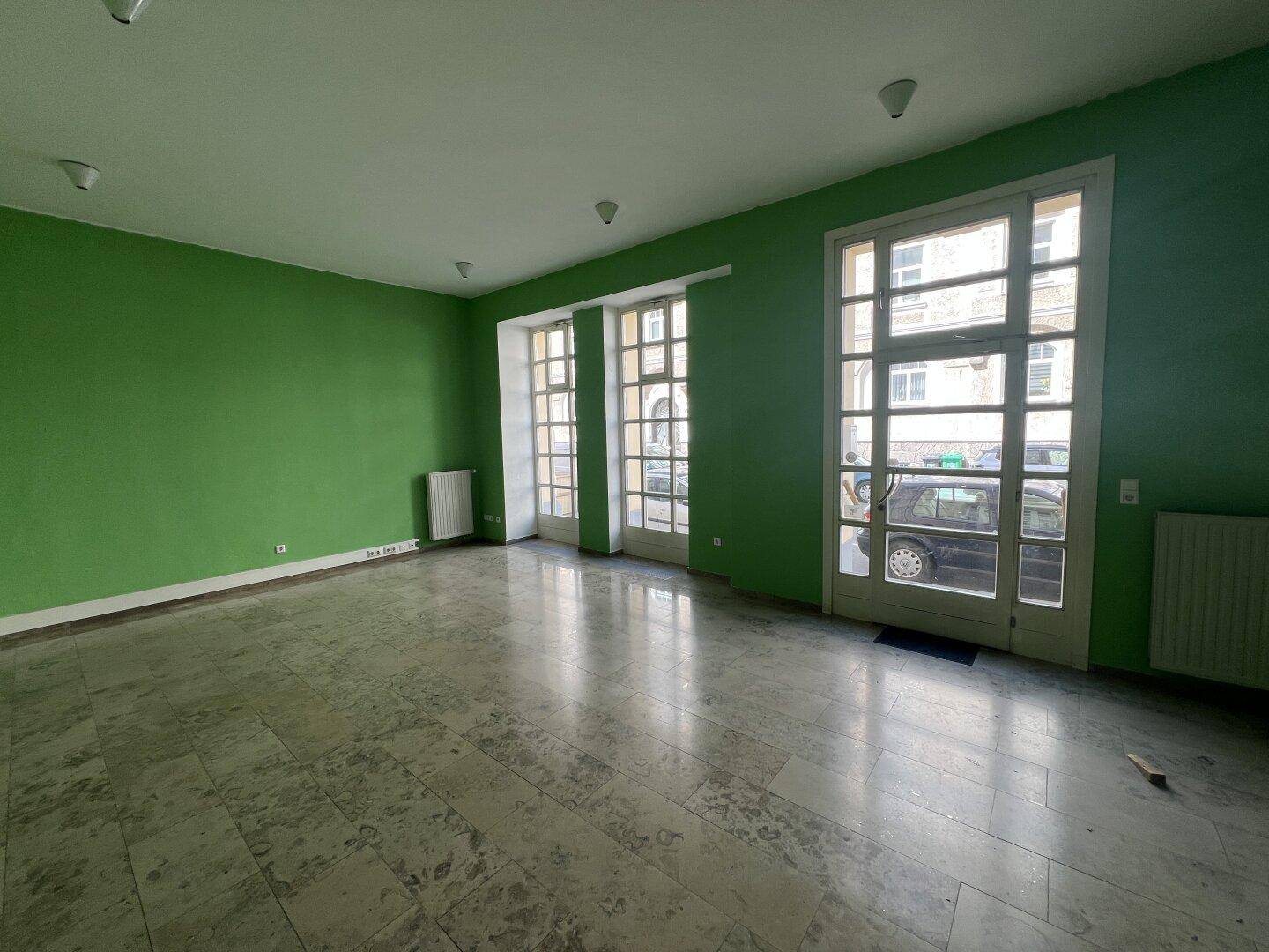 Leerer Raum mit auffällig grünen Wänden und großen Fenstertüren zur Straße hin.