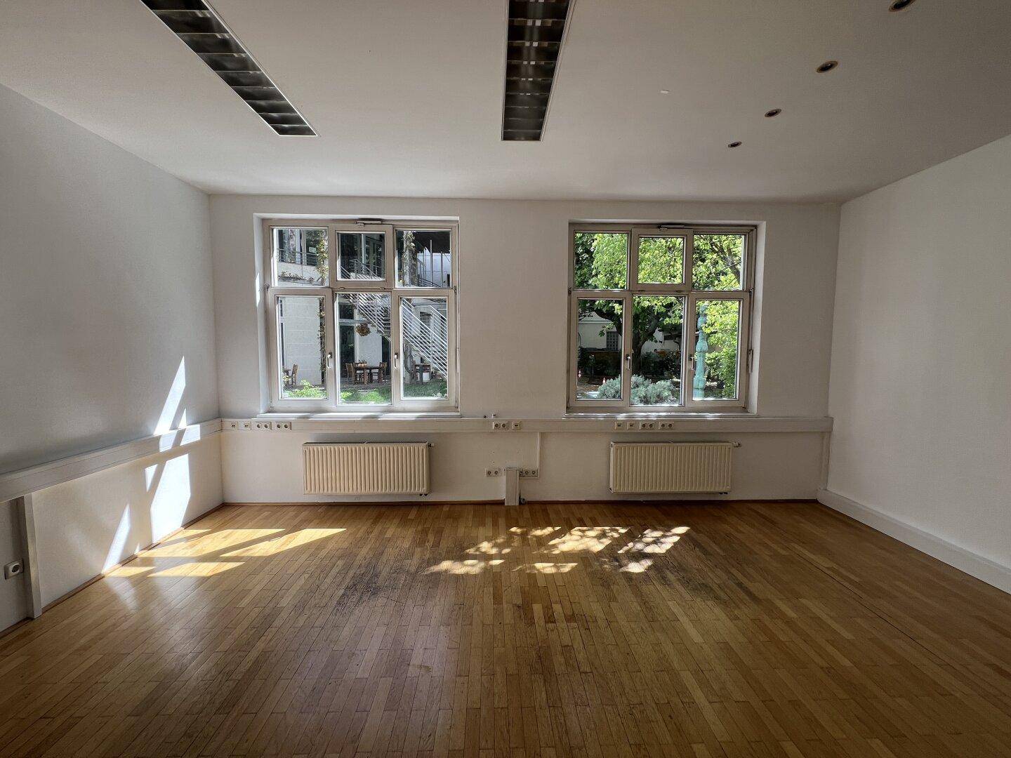 Heller, leerer Raum mit Holzboden und großen Fenstern, die viel Tageslicht hereinlassen.