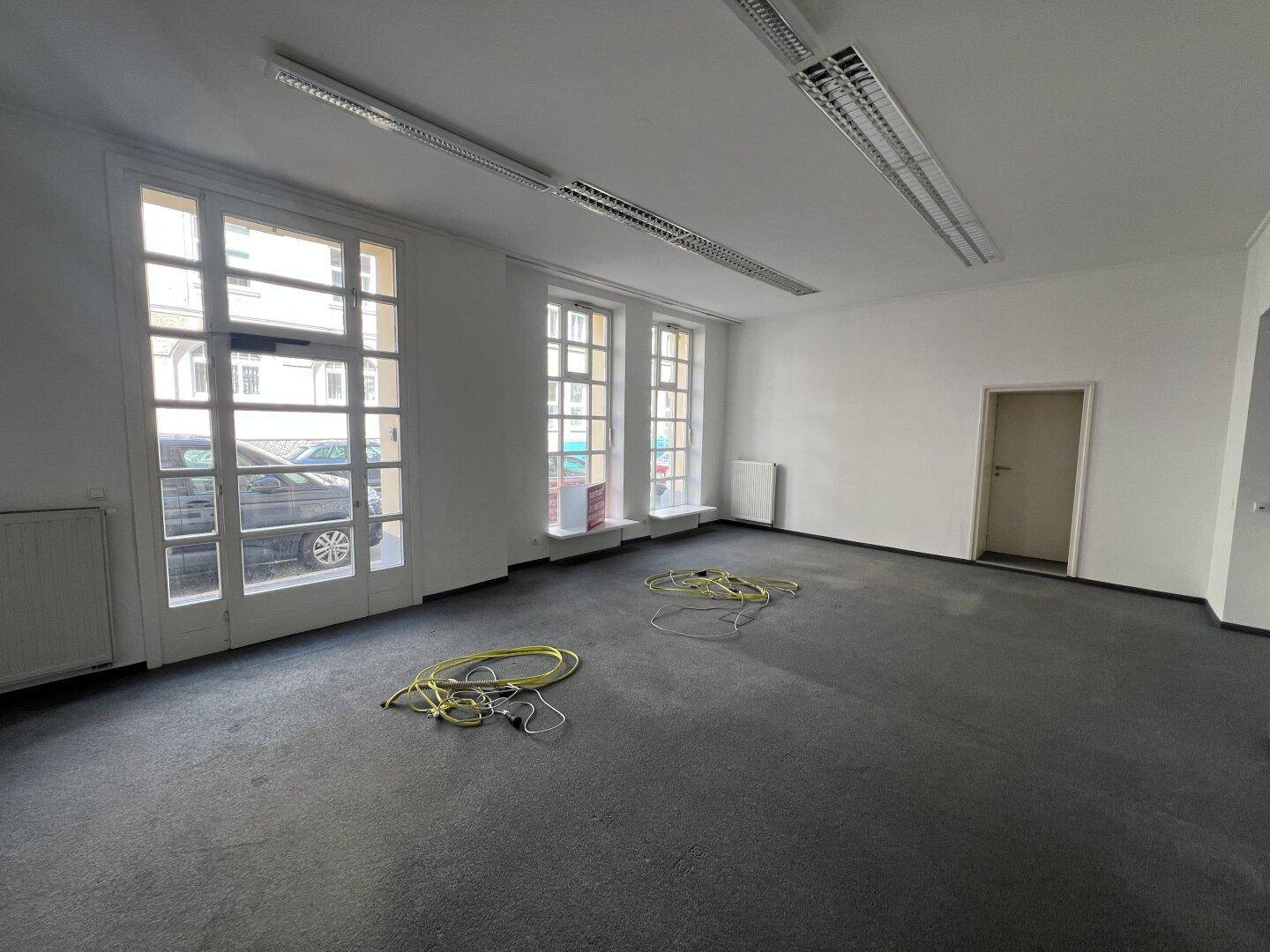 Geräumiger, leerer Geschäftsraum mit Teppichboden und großen Fenstern zur Straße.