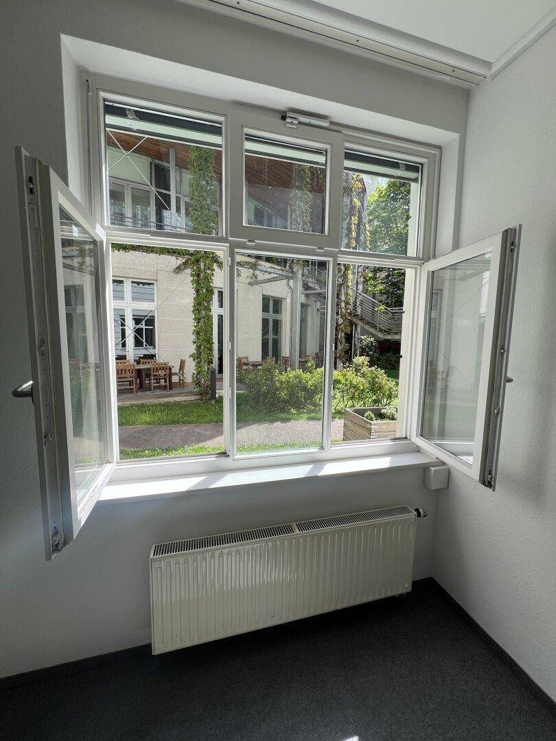 Fensterblick aus einem Büro in den grünen Innenhof mit Sitzgelegenheiten und Bäumen.