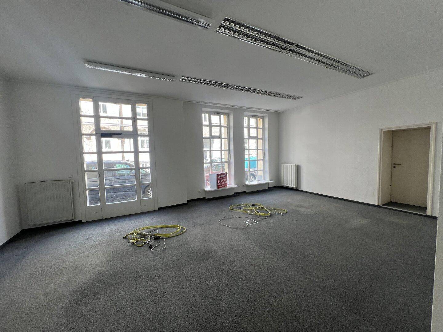 Großer, leerer Geschäftsraum mit Teppichboden und hohen Fenstern zur Straße.
