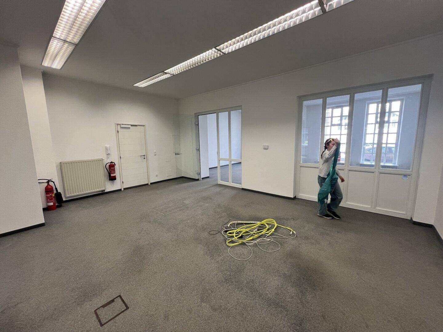 Geräumiger Geschäftsraum mit Teppichboden, Glastüren und einem Feuerlöscher an der Wand.