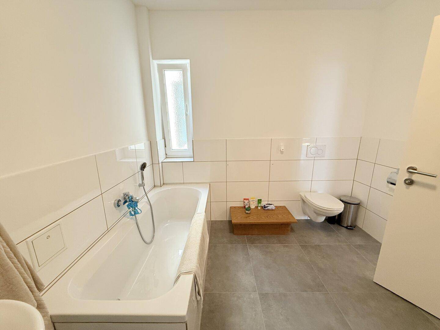 Modernes Badezimmer mit Badewanne, Wand-WC, grauen Fliesen und Fenster.