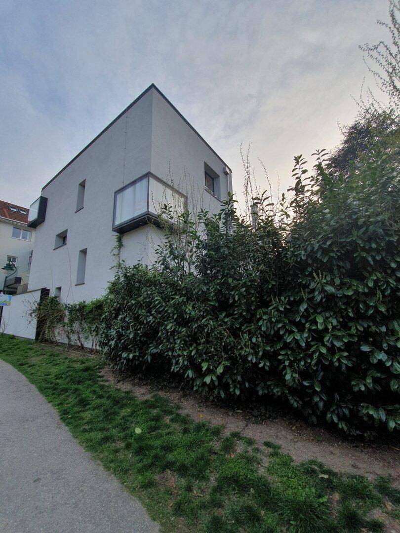 Modernes Gebäude mit weißer Fassade, umgeben von dichten grünen Hecken und Bäumen.