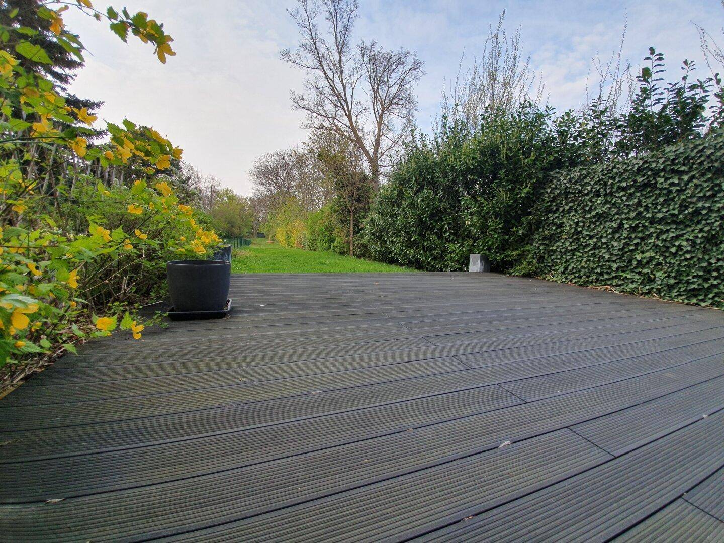Terrasse mit Holzboden und Zugang zu einem grünen Gartenbereich mit Hecken.