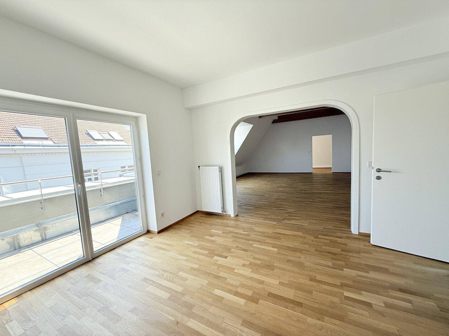 Heller Wohnraum mit Parkettboden, großem Terrassenzugang und offenem Durchgang zu weiteren Zimmern.