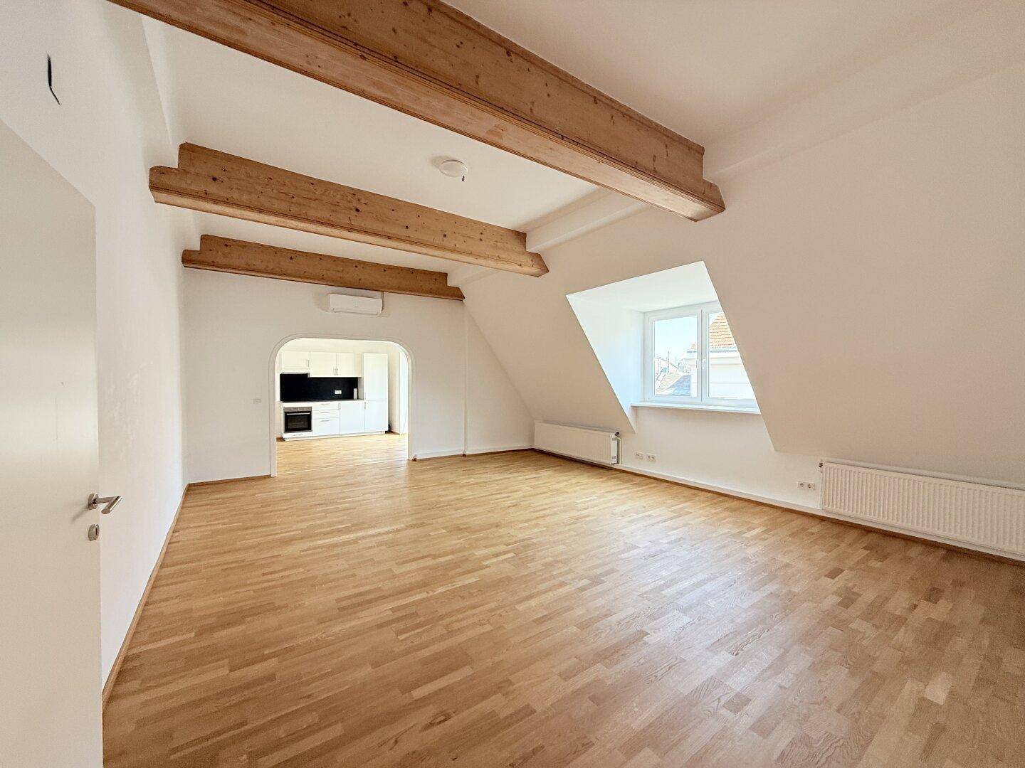 Geräumiger Wohnbereich mit Holzbalken, Parkettboden und Zugang zu einer modernen Küche.
