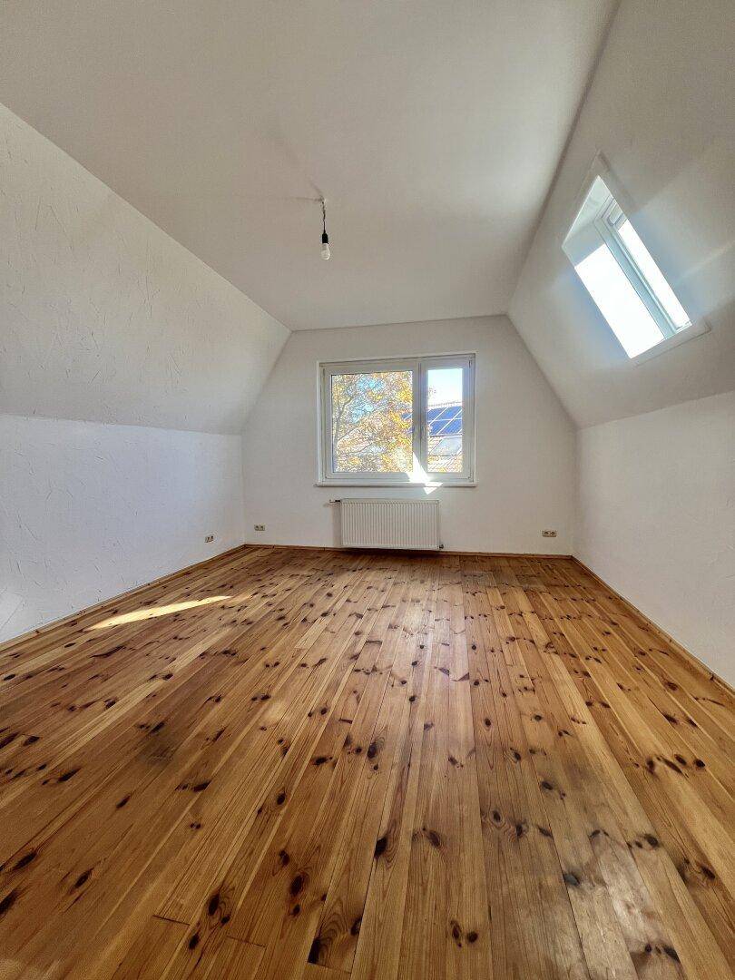 Heller, leerer Raum mit Holzboden, Dachschräge und großem Fenster.