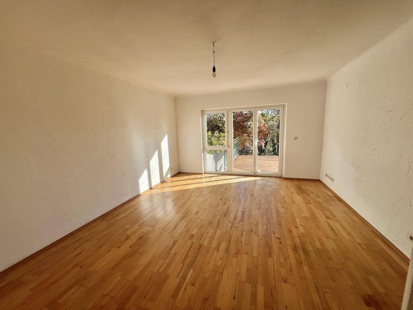 Geräumiges, leeres Wohnzimmer mit Holzboden und großen Fenstern, die viel Licht hereinlassen.
