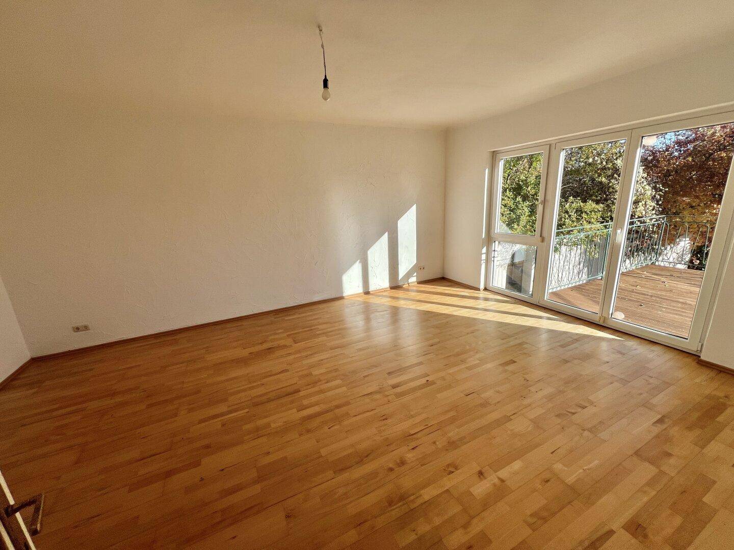 Geräumiges, leeres Wohnzimmer mit Holzboden und großen Fenstern, die viel Licht hereinlassen.