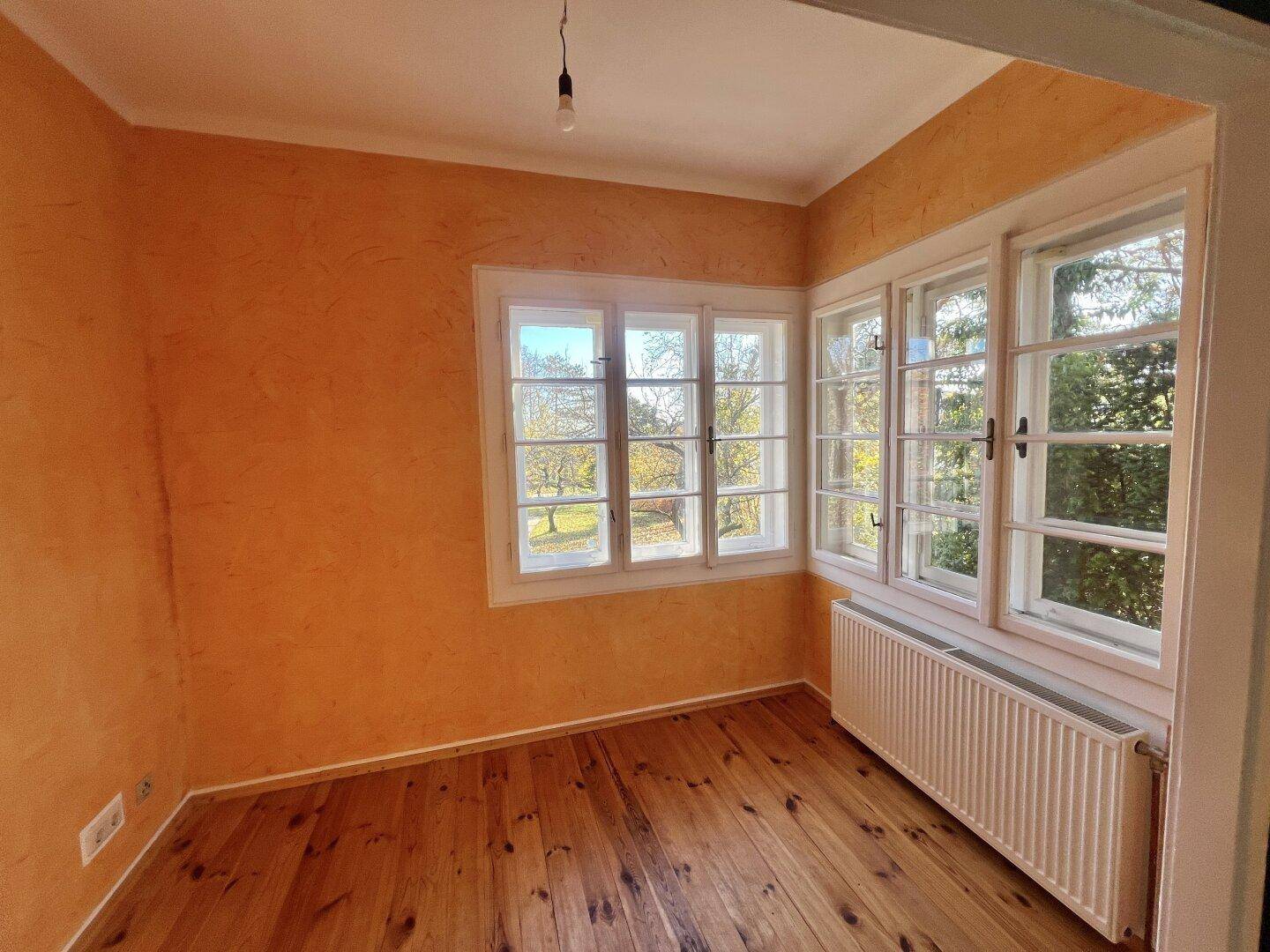 Heller Raum mit orangefarbenen Wänden, Holzboden und vielen Fenstern mit Blick ins Grüne.