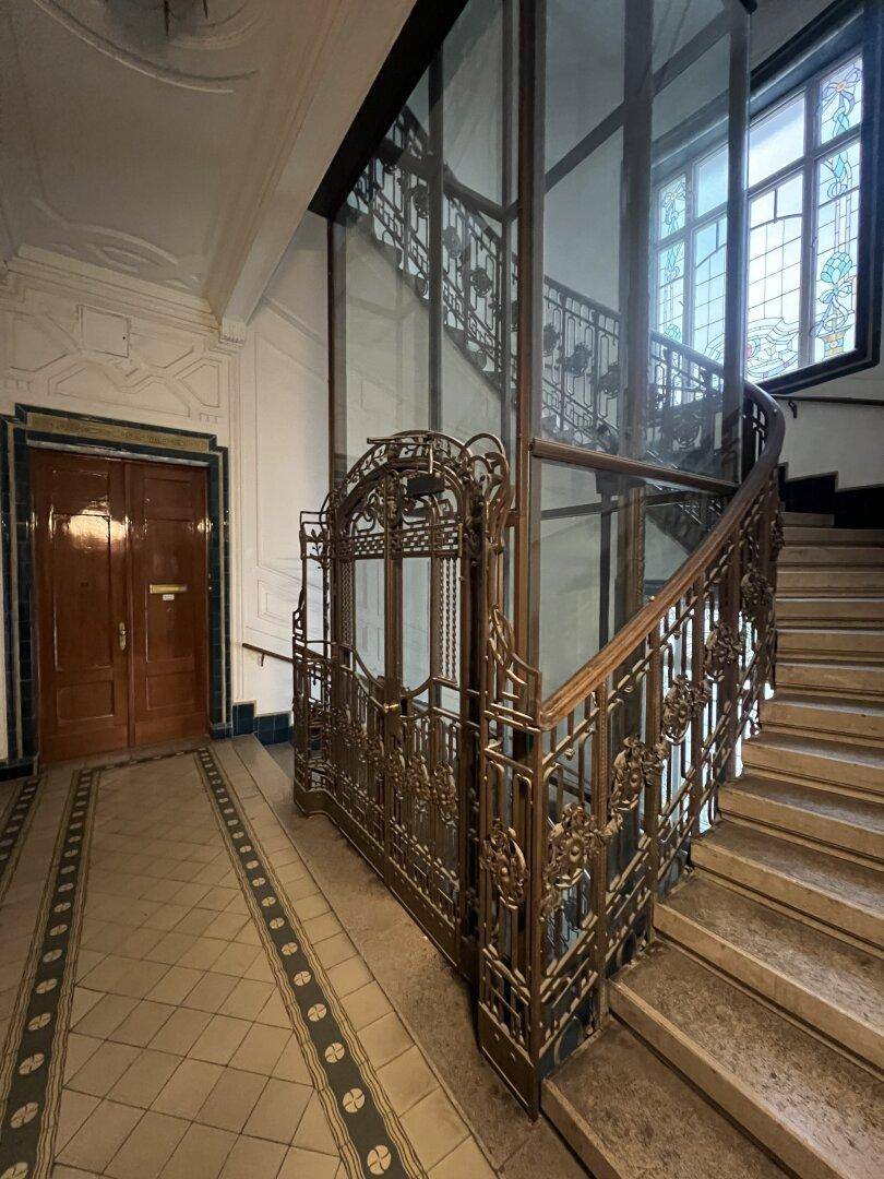 Historisches Treppenhaus mit schmiedeeisernem Geländer und einem modernen Glasaufzug.