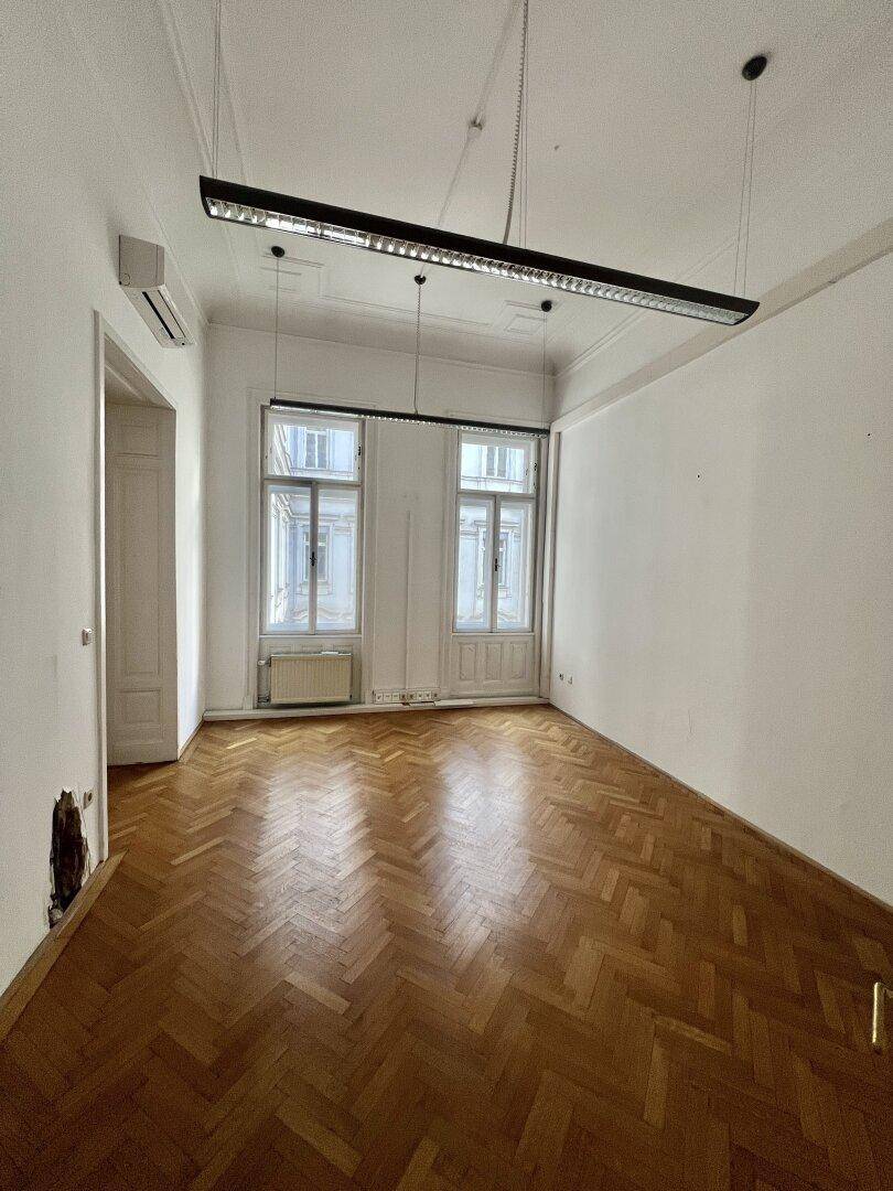 Leerer Raum mit Fischgrätparkett, hohen Decken und zwei großen Fenstern, bereit zur individuellen Gestaltung.