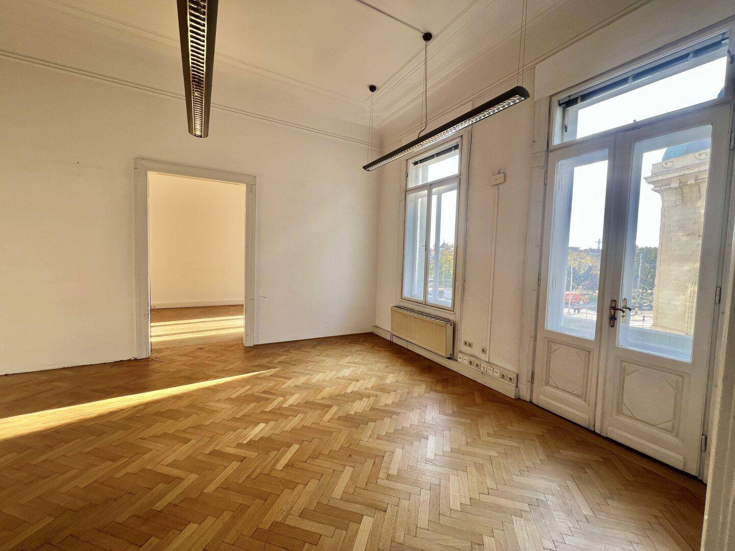 Heller, leerer Raum mit Fischgrätparkett, hohen Fenstern und Blick auf die Stadt, ideal für ein Büro.