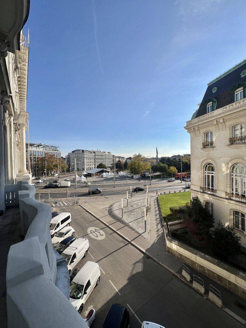 Blick vom Balkon auf eine belebte Stadtstraße mit historischen Gebäuden und blauem Himmel.