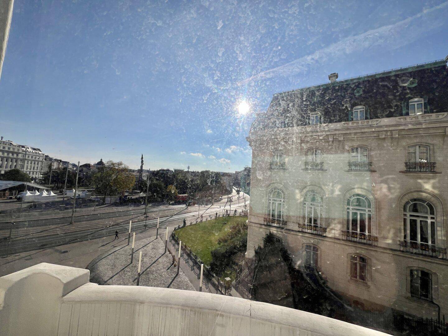 Blick durch ein Fenster auf eine belebte Stadtstraße mit Gebäuden und blauem Himmel.