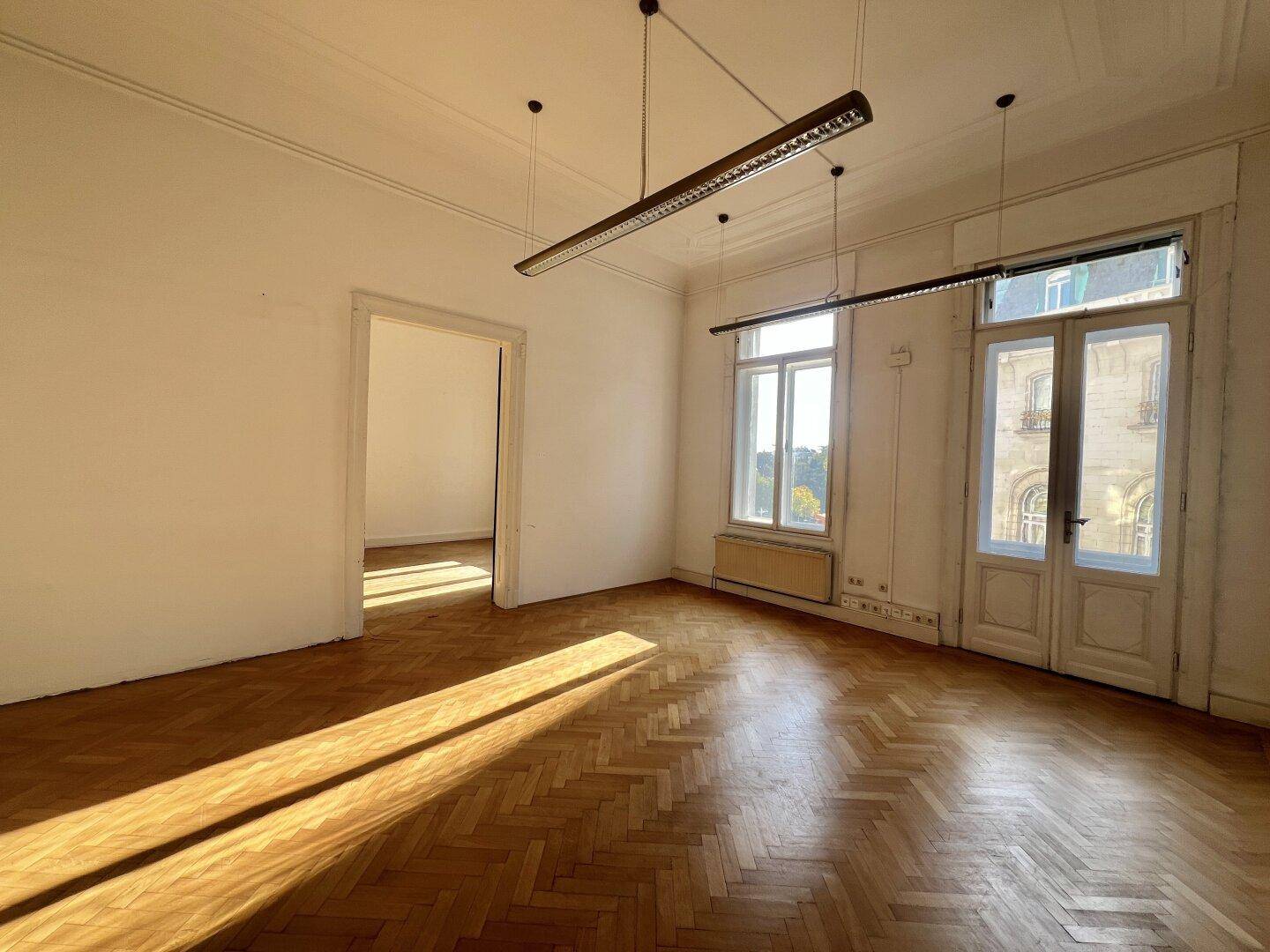 Sonnendurchfluteter, leerer Raum mit Fischgrätparkett, hohen Decken und großen Fenstern, ideal für ein Büro.