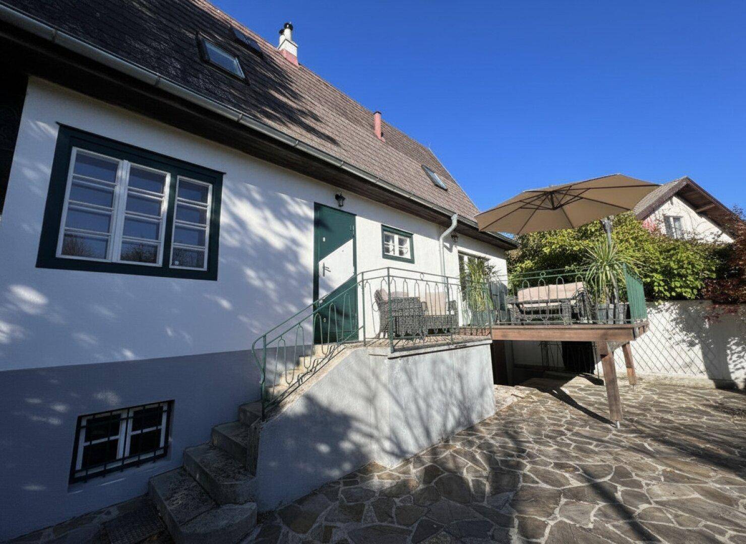 Hausfassade mit erhöhter Terrasse, Sitzgelegenheiten und Sonnenschirm, umgeben von Grün.