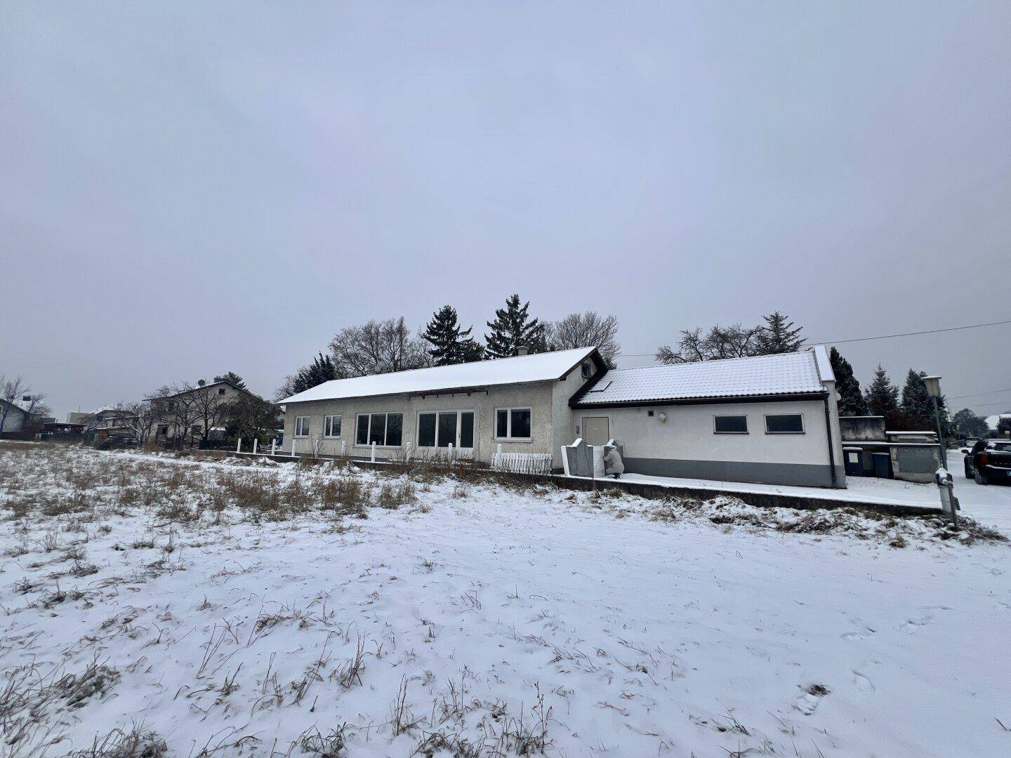 Hausfassade im Winter mit schneebedecktem Boden und Bäumen unter einem grauen Himmel.