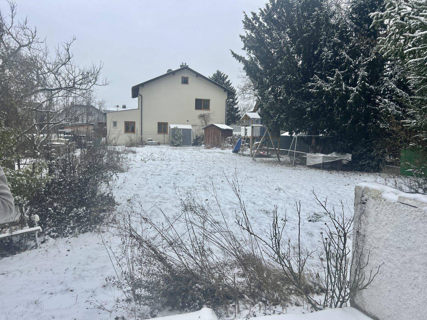 Verschneite Gartenlandschaft mit Bäumen, einem Spielplatz und dem Haus im Hintergrund.