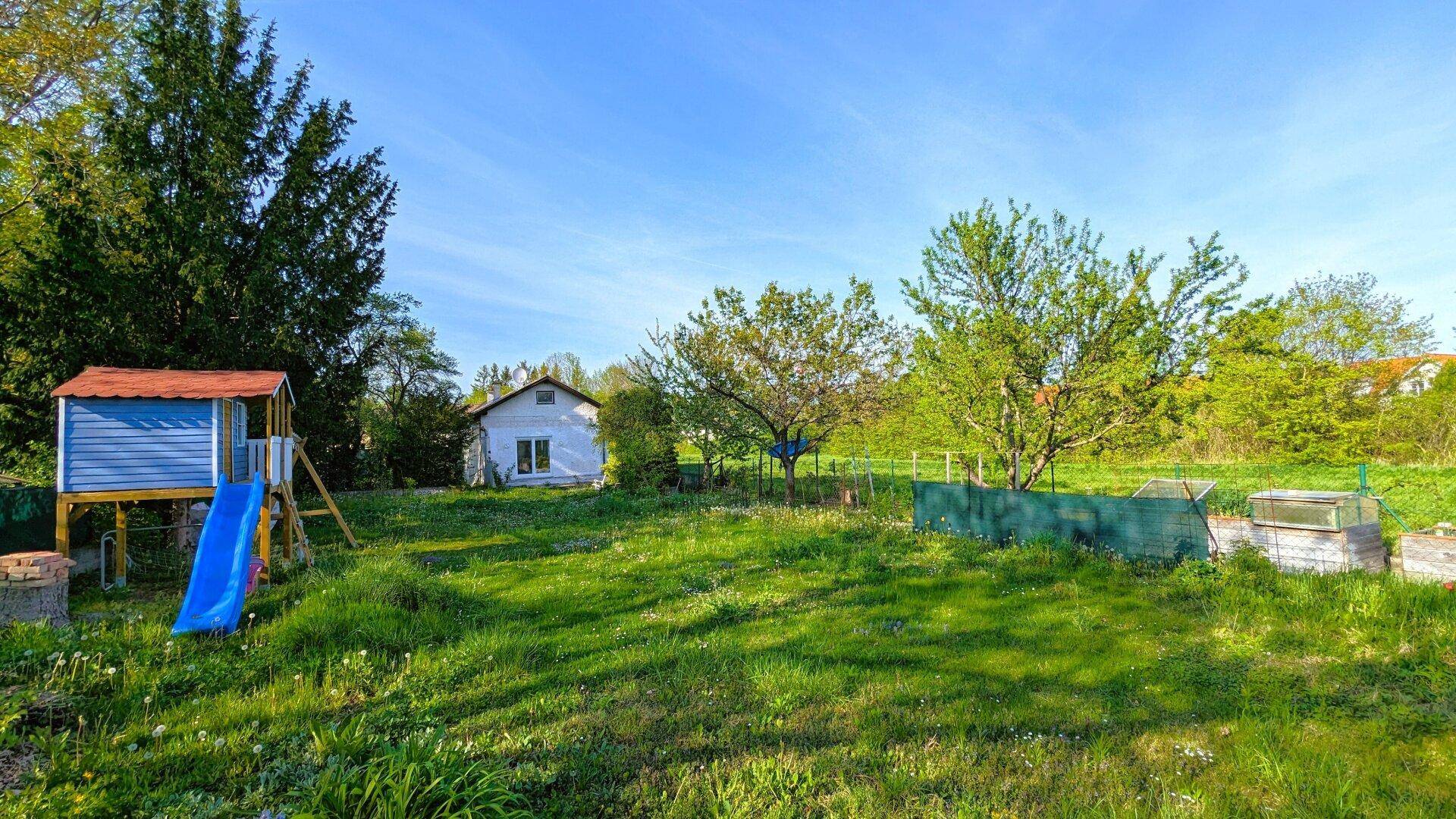 Sonniger Garten mit Spielplatz, Bäumen und einer weitläufigen grünen Rasenfläche.
