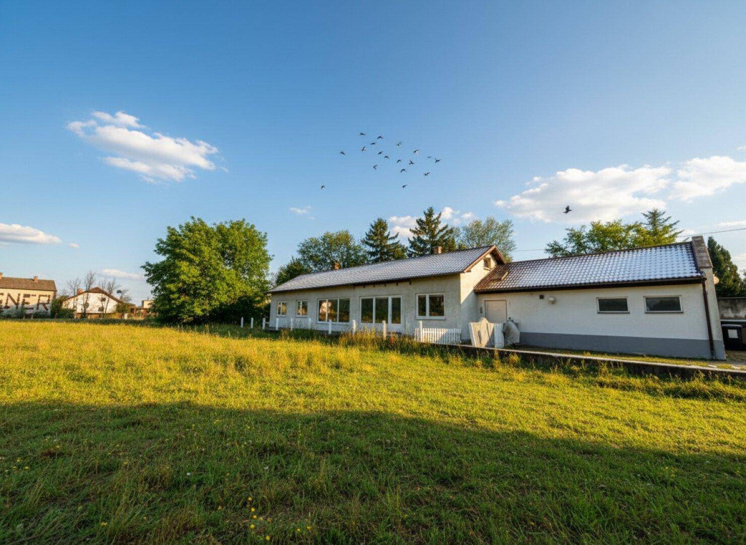 Hausfassade mit weitläufiger Rasenfläche und Bäumen unter einem strahlend blauen Himmel.