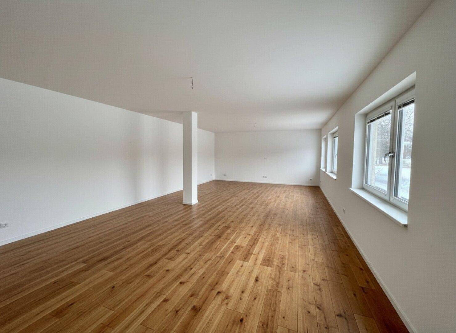 Geräumiger, leerer Raum mit hellem Holzboden, weißen Wänden und großen Fenstern.