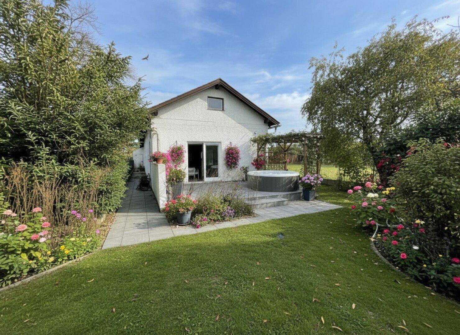 Gepflegter Gartenbereich mit Terrasse, Whirlpool und blühenden Blumen unter blauem Himmel.