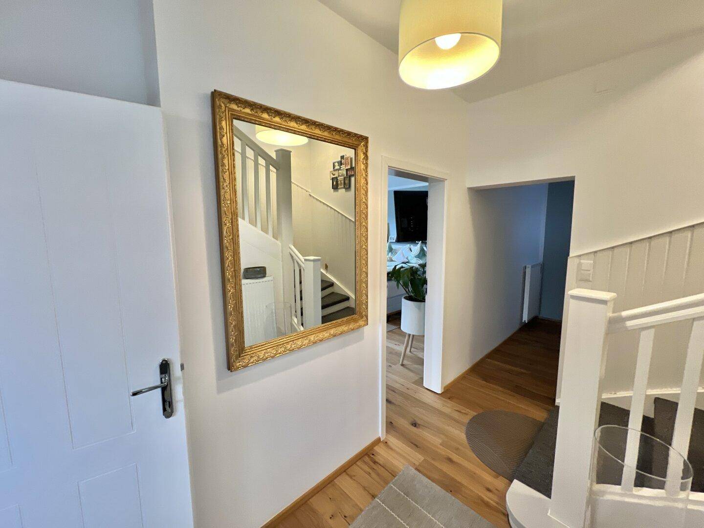 Heller Flur mit Holzboden und Treppenaufgang, geschmückt mit einem goldenen Spiegel.