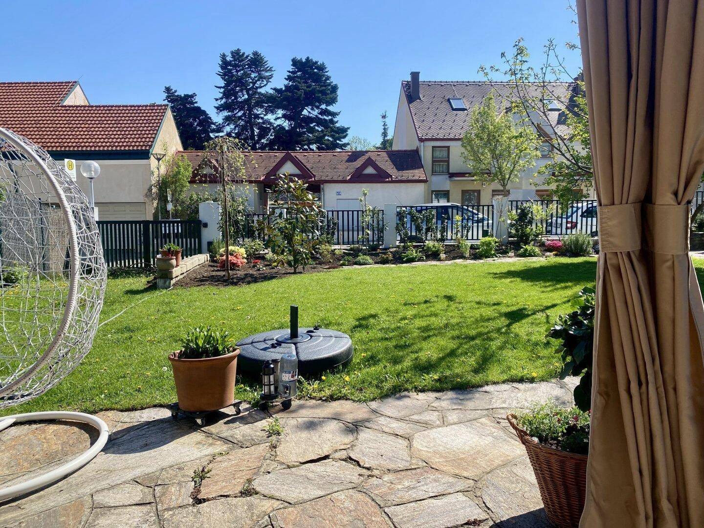 Sonnige Gartenecke mit Hängesessel und Blick auf die umliegenden Häuser.