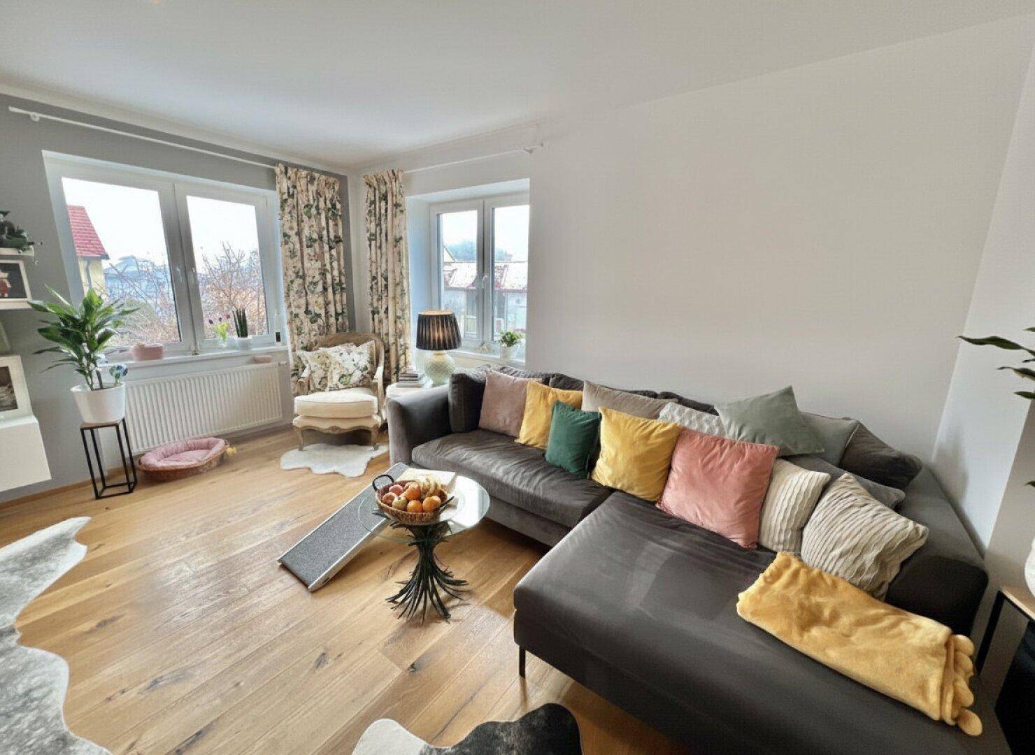 Helles Wohnzimmer mit großem Ecksofa, Holzboden und Fenstern mit Blick ins Grüne.