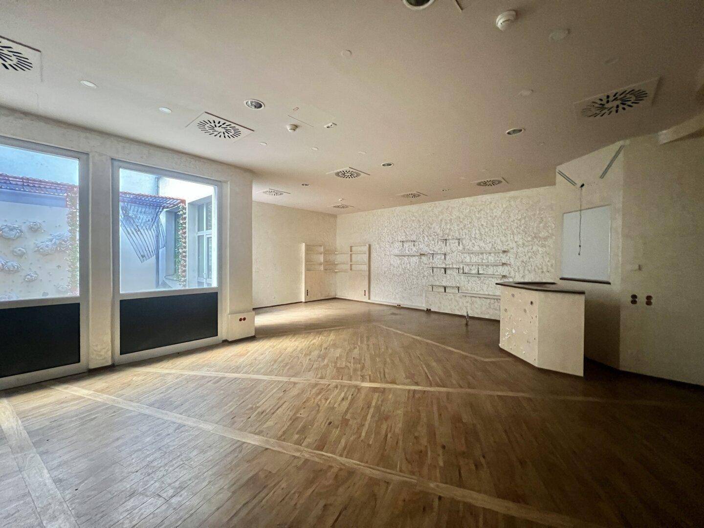 Heller Gewerberaum mit Holzboden, großen Fenstern und strukturierten Wänden, ideal für Laden oder Büro.