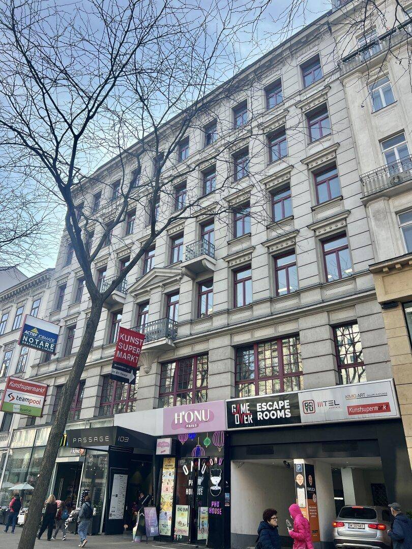 Mehrstöckiges Gebäude mit klassischer Fassade, Geschäften im Erdgeschoss und Balkonen, an einer städtischen Straße.