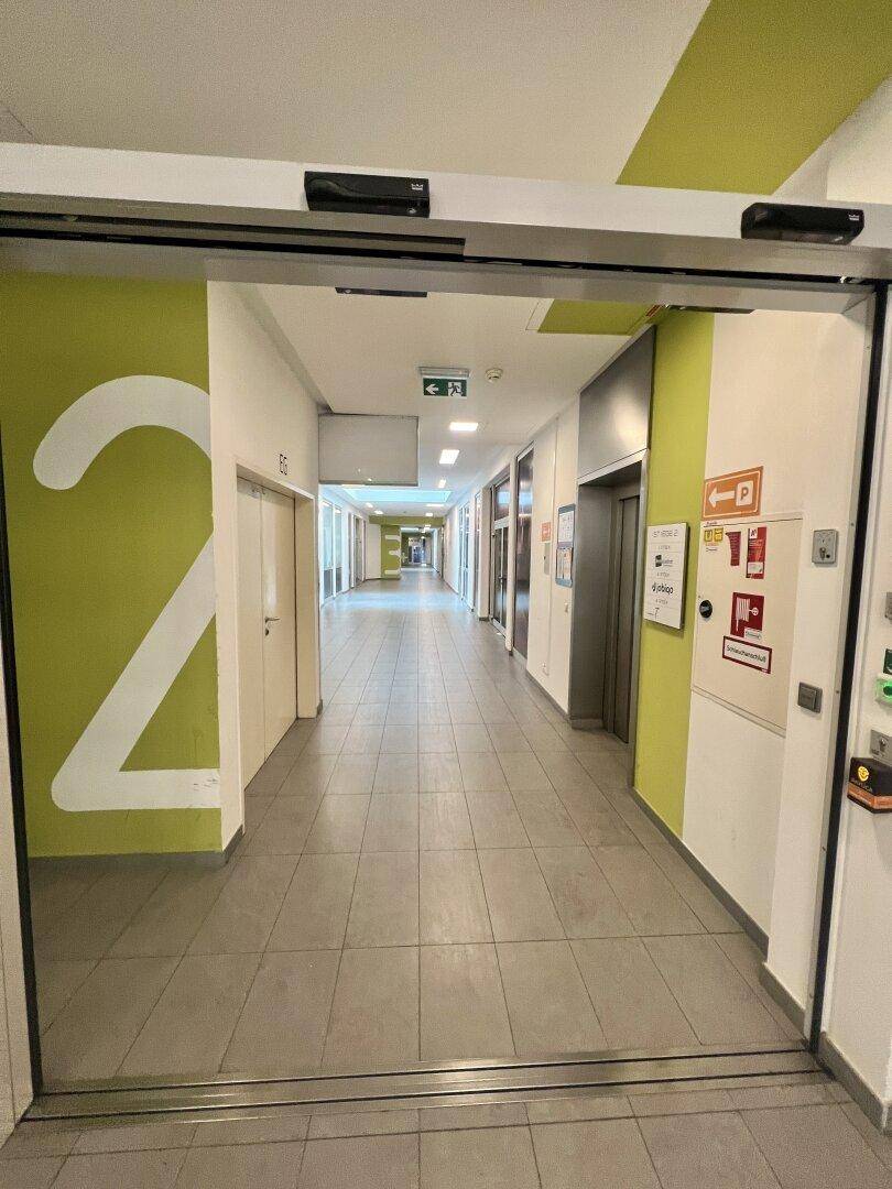 Heller Flur mit Fliesenboden und grünen Akzentwänden, führt zu Geschäften und einem Aufzug.