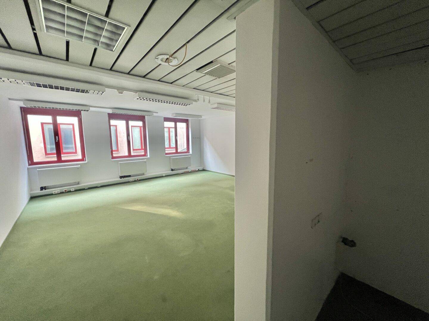 Leerer Büroraum mit grüner Teppichboden, weißen Wänden und Blick auf einen angrenzenden Bereich.