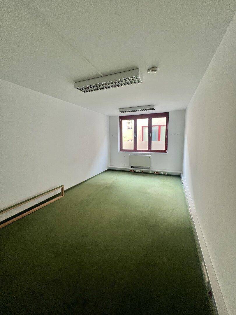 Leerer Büroraum mit grüner Teppichboden, einem Fenster und weißen Wänden, bereit zur Einrichtung.