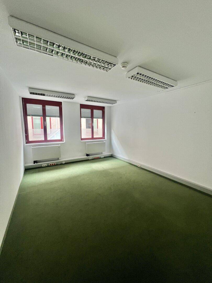 Leerer Büroraum mit grüner Teppichboden, zwei Fenstern und weißen Wänden, ideal für Einzelbüro.