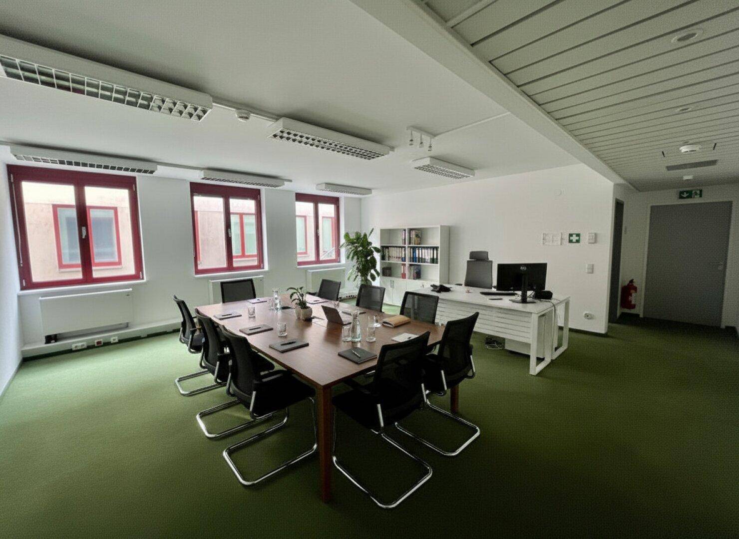 Großer Konferenzraum mit einem langen Holztisch, vielen Stühlen und Fenstern mit rotem Rahmen.