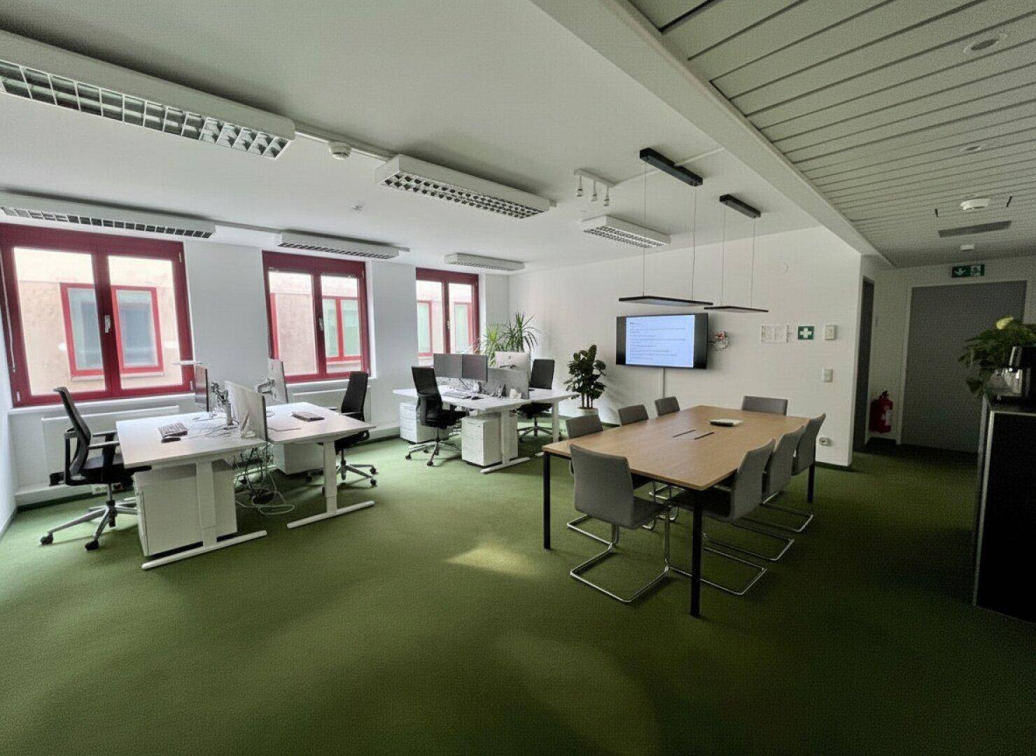 Großes offenes Büro mit mehreren Arbeitsplätzen, einem Konferenztisch und grüner Teppichboden.