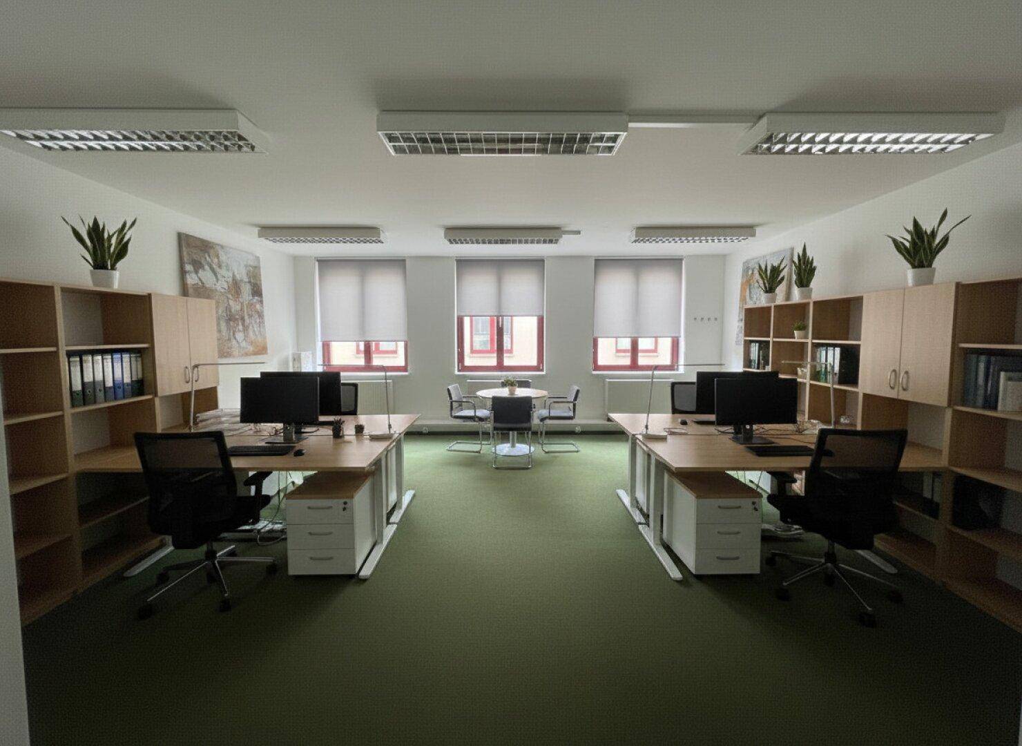 Geräumiges Büro mit zwei Reihen von Arbeitsplätzen, Regalen und Fenstern mit Jalousien.