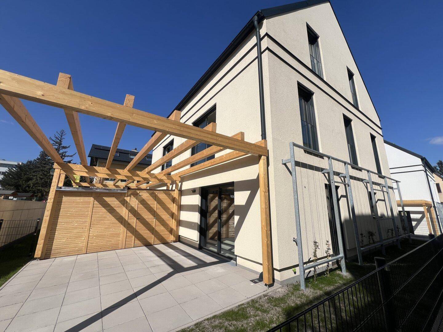Moderne Hausfassade mit großer gepflasterter Terrasse, Holzpergola und kleinem Rasenbereich.