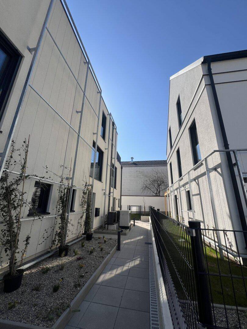 Schmaler gepflasterter Weg zwischen zwei modernen Gebäuden mit bepflanzten Kiesbeeten.